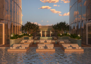 A luxurious outdoor swimming pool area at sunset, featuring cushioned sun loungers, stacked towels, potted plants, lanterns, and two modern glass buildings reflecting the sky with clouds.